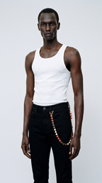 A male model wearing a white ribbed tank top and black jeans with a decorative red and gold wallet chain hanging from the waistband, photographed against a light gray studio background.