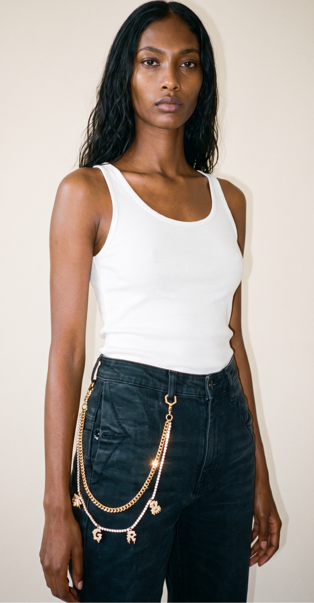 A woman modeling a white ribbed tank top and dark jeans, accessorized with a gold wallet chain belt featuring letter charms, spelling out bee great